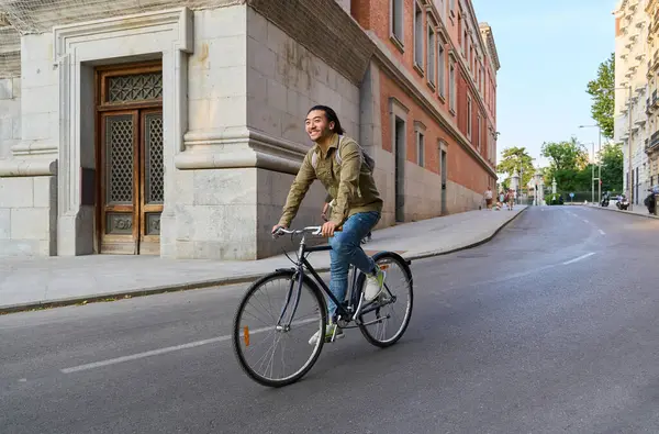 Güneşli bir şehir caddesinde bisiklet sürerken gülümseyen genç erkek öğrenci canlı şehir atmosferinin ve sağlıklı yaşam tarzının tadını çıkarıyor.
