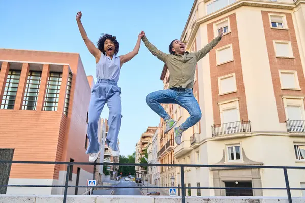 Neşeli erkek ve kadın şehirde sevinçten havalara uçuyor, başarılarını kollarını kaldırıp el ele tutuşarak kutluyorlar.