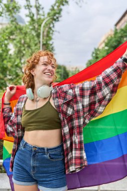 Boynunda kulaklıkları olan mutlu bir kızıl kadın, ekose gömlek giyiyor ve gay onur yürüyüşünde gökkuşağı bayrağı taşıyor.