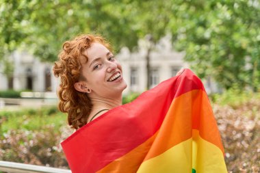 Genç kızıl saçlı kadın gururla gülümserken neşeli bir gay geçit töreninde elinde gökkuşağı bayrağı sallıyor.