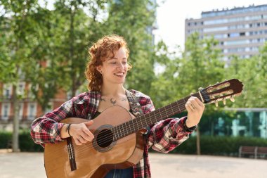 Parkta İspanyol gitarı çalan bir sokak sanatçısı, güneşli bir günde müziğin ve canlı şehir yaşamının neşesini kucaklıyor.