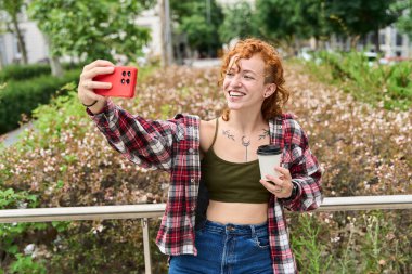 Dövmeli, kızıl saçlı, gülümseyen ve cep telefonuyla selfie çeken genç bir kadın şehir parkında tek kullanımlık kahve fincanı tutuyor.