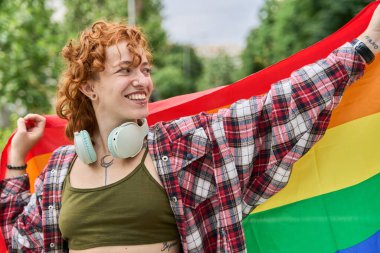 Boynunda kulaklıkları ve elinde gökkuşağı bayrağı olan mutlu genç bir kadın lgbtq 'ı kutluyor.