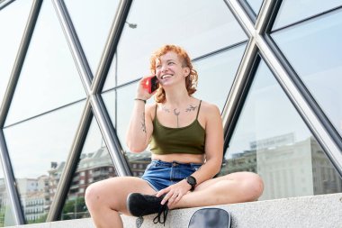 Moda dövmeli mutlu bir kadın cep telefonunu kullanıyor, arkasında modern cam bir binayla caddede oturuyor.