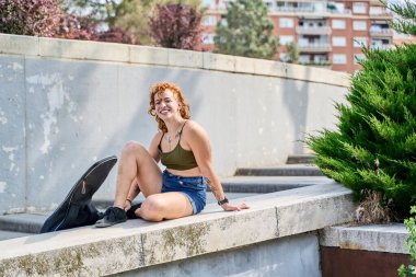 Şehir ortamında beton duvarda oturan mutlu kızıl kaykaycı, kaykay antrenmanından sonra mola veriyor.