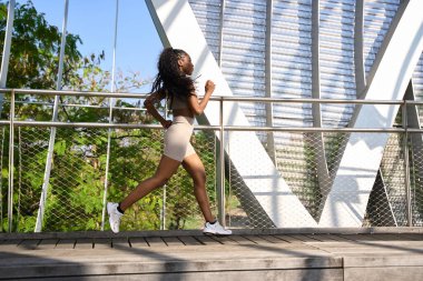 Şehrin modern bir köprüsünde koşan sağlıklı bir kadın, açık hava çalışmasının enerjisini kucaklıyor ve canlı şehir manzarasının tadını çıkarıyor.