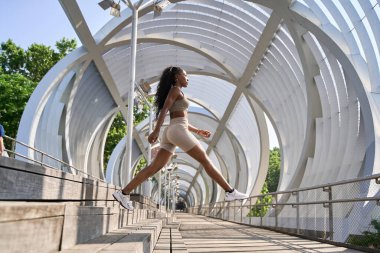 Güneşli bir günde, kentteki modern beyaz metal bir köprünün merdivenlerinden atlayan esneme egzersizleri yapan zinde bir zenci kadın.
