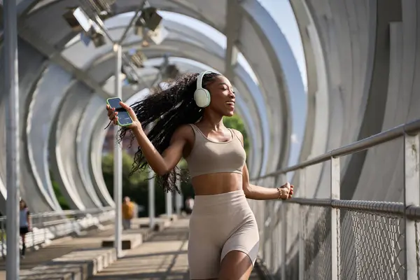 Şehirdeki bir köprüde kablosuz kulaklıkla müzik dinleyen mutlu sportif kadın.