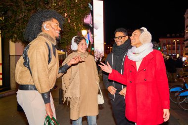 Dört arkadaş kış akşamı gezintisinin keyfini çıkarıyorlar, aydınlatılmış şehir sokaklarında gezinirken canlı sohbetler yapıyorlar.