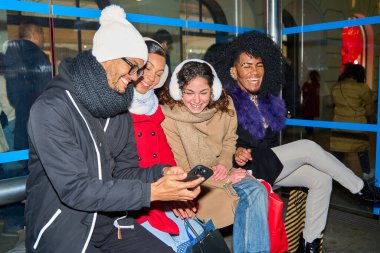 Dört neşeli çoklu etnik arkadaş akıllı telefon kullanıyor ve noel alışverişine ara verirken birlikte gülüyorlar.