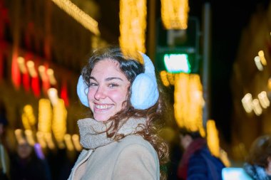 Neşeli genç kadın kulaklık takarken gülümsüyor, parıldayan ışıklar altında hareketli bir Noel pazarının neşeli atmosferinin tadını çıkarıyor.