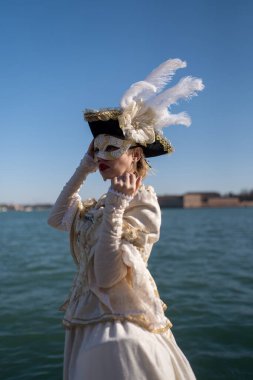 Venetian carnival participant in an ornate ivory costume with a feathered hat, elegantly covering her mask against a scenic waterfront backdrop. clipart