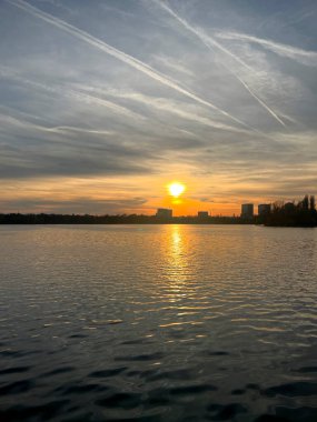 Gün batımına yakın, gökyüzünde uçak izleri var ve güneş suya yansıyor.