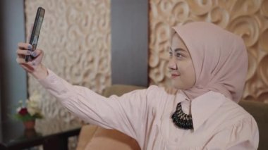 Modern Muslim Woman on Hijab Southeast Asian - Smiling, Pose, Taking Selfie Photo with her Smartphone while Sitting on Couch in The Living Room