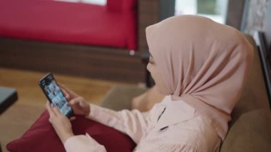 Modern Muslim Woman on Hijab Southeast Asian - Smiling, Looking and Scrolling her Smartphone while Sitting on Couch in The Living Room