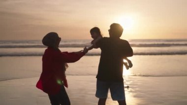Happy Modern Muslim Southeast Asian Indonesian Family Enjoying Sunset Together on The Beach. Silhouette of Father Mother and Child in Slow Motion.