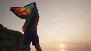 Koyu tenli Güneydoğu Asya LGBT Gay Adam Gün batımında Gökkuşağı Gurur Bayraklarıyla Sahilde Dans Ediyor