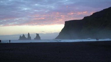 Black Sand Beach Vik, İzlanda 'da gün batımı. Reynisdrangar kayalıkları ve deniz yığınları. Vik 'in çevresindeki uçurumların ve taş yığınlarının manzarası. İnsanlar sahilde dalgalarla koşup oynuyorlar..