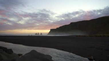Black Sand Beach Vik, İzlanda 'da gün batımı. Reynisdrangar kayalıkları ve deniz yığınları. Vik 'in çevresindeki uçurumların ve taş yığınlarının manzarası. İnsanlar sahilde dalgalarla koşup oynuyorlar..