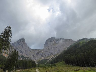An idyllic narrow pathway leading to the Alpine Valley in the region of Eisenerz, Austria. There are thick clouds above the high mountain chains. Dense forest at the foothill. Stony and sharp slopes