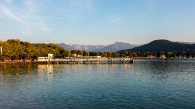 A panoramic capture of the pier on the Woerthersee lake in Austria. In the back there are high Alpine chains. The shore of the lake is overgrown with dense forest. Serenity and calmness. Golden hour.