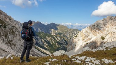 A man with a big hiking backpack admiring the view on a stony valley in Italian Dolomites. There are high and sharp mountains around. Remote and raw landscape. Sunny day. Freedom and serenity