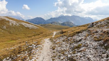A narrow pathway along a high plateau in Italian Dolomites. There are golden mountain slopes around the pathway. High mountain chains in the back. Desolated and remote landscape. A bit of overcast