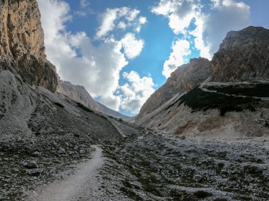 A panoramic view on a stony valley in Italian Dolomites. There are high and sharp mountains around. A narrow pathway leading through the stones. Remote and raw landscape. Sunny day. A bit of overcast
