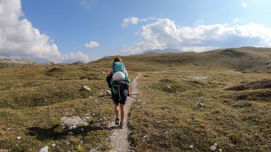 A woman with a big hiking backpack hiking on a plateau in high Italian Dolomites. There are golden mountain slopes around the pathway. High and sharp mountains around. Remote and raw area. Calmness