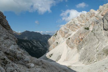 A panoramic view on a stony valley in Italian Dolomites. There are high and sharp mountains around. A narrow pathway leading through the stones. Remote and raw landscape. Sunny day. A bit of overcast