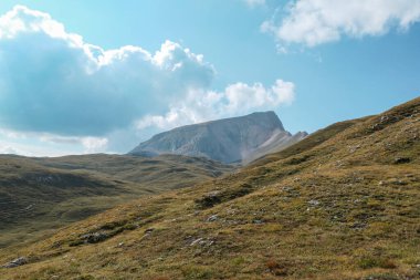 A panoramic view on a high plateau in Italian Dolomites. There are high mountains around. The area is partially overgrown with grass and partially covered with stones and pebbles. Remote and desolate