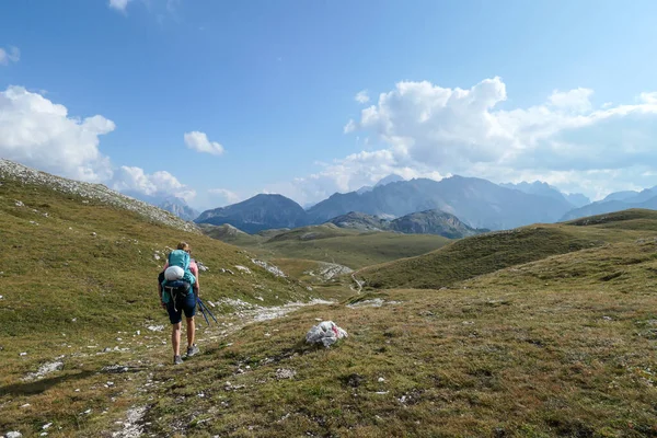 A woman with a big hiking backpack hiking on a plateau in high Italian Dolomites. There are golden mountain slopes around the pathway. High and sharp mountains around. Remote and raw area. Calmness