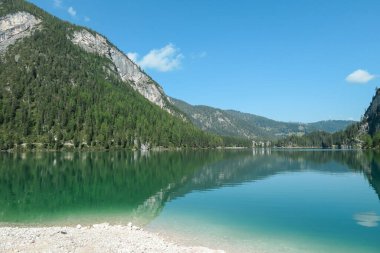A panoramic view on the Pragser Wildsee, a lake in South Tyrolean Dolomites. High mountain chains around the lake. The sky and mountains are reflecting in the lake. Dense forest at the shore. Serenity
