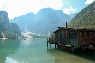 A small wooden house build on pillars above the Pragser Wildsee, a lake in South Tyrolean Dolomites. High mountain chains around the lake. Dense forest at the shore. Autumn vibe. Serenity and calmness