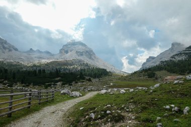 A panoramic view on a valley with a gravelled road along it in Italian Dolomites. There is a wooden fence along the road. Sharp and stony mountain chains in front. Few boulders on the green meadow.