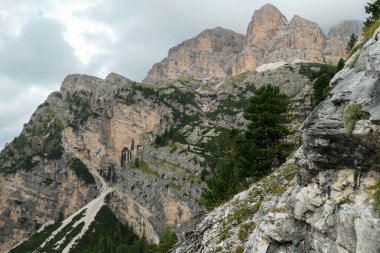 A view on a sharp and stony mountain wall in Italian Dolomites. The massive mountain is surrounded by clouds. Lower parts overgrown with trees. Remote and desolate landscape. Silence before the storm