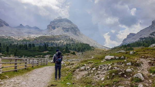 A man with a big hiking backpack hiking along a gravelled road in Italian Dolomites. There is a wooden fence along the road. Sharp and stony mountain chains in front. Few boulders on the green meadow.