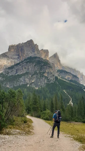A man with a hiking backpack hiking on a gravelled pathway in high Italian Dolomites. There are a few trees on the lower parts, and steep, sharp mountain chain in the back. Discovering and exploring
