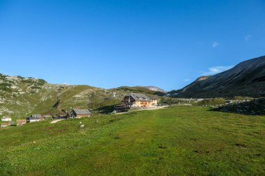 A small Sennes cottage, located on a lush green meadow in high Italian Dolomites. The wooden cottage is surrounded by high mountains. There are a few smaller cottages around. Bright and sunny day.
