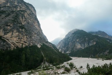 A panoramic view on a vast valley in Italian Dolomites. The bottom of the valley is very wide, covered with small pebbles. Sharp and tall mountains around, shrouded with thick clouds. Coming storm