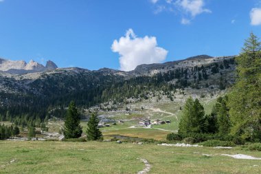 A panoramic view on the valley in Italian Dolomites. Dense forest overgrows the mountains slopes. Lush green meadow in front. High mountain chains in the back. Spring in the mountains. Sunny day.