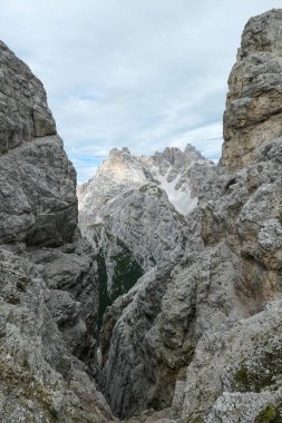 A sneak-peak on a high and distant mountain in Italian Dolomites. The mountain is visible between two rocky and steep slopes in front. Playing hide and seek. Raw and desolate landscape. Overcast