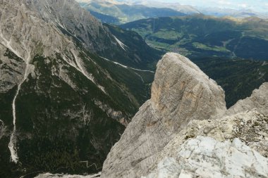 A panoramic view on a valley in Italian Dolomites. There is a massive mountain on the side, with very steep and sharp slopes. In the back there are smaller mountain chains, overgrown with green plants