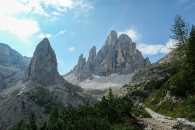 A panoramic view on Italian Dolomites. There are many high and sharp peak in front, with many landslides. Dangerous climbing. There are few trees and bushes on the side. Raw and unspoiled landscape
