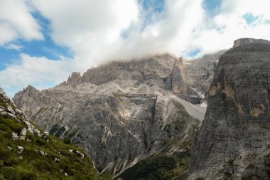 A panoramic view on Italian Dolomites. There are many high and sharp peaks covered with light clouds. Dangerous landslides. The slopes are overgrown with moss. Raw and unspoiled landscape.