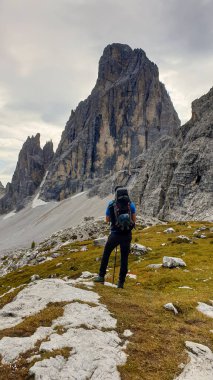 Man with big backpack and sticks, hiking in high Italian Dolomites. There are many sharp peaks in front of him. He is standing on a big boulder. Lots of lose stones and landslides. Sunny day. Outdoor