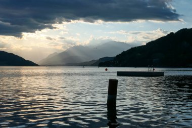 A panoramic view on the Millstaettersee lake from its shore. The lake is surrounded by high mountains. A wooden platform drifting on its surface. Thick and dark clouds above. Golden hour. Calmness