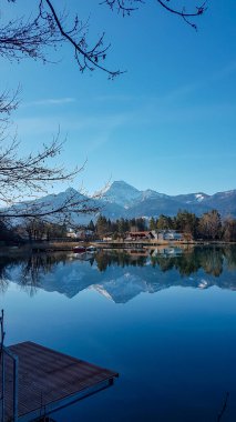 Panoramic view on the Faaker lake in Austrian Alps. The lake is surrounded by high mountains. A small wooden pier. Calm surface of the lake reflects the surrounding. Sweet flag at the shore. Blue sky