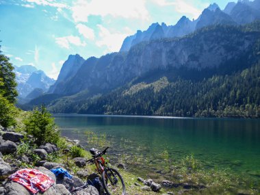 Gosau Gölü kıyısında turuncu bir bisiklet, Avusturya Alplerinde Dachstein buzulu. Göl yüksek dağlarla çevrilidir, yüksek ağaçlarla kaplıdır. Güneş yüzeye yansıyor. Idyll.