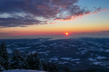 A sunrise seen from snow-capped peak of Schoeckl in Austrian Alps. The sky is bursting with orange and pink. Distant view on Graz, shrouded with fog. Winter wonderland. Day break. Calmness